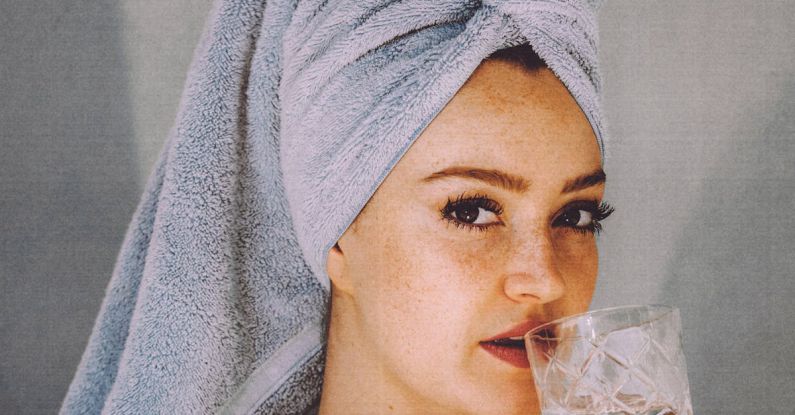 Clean Care - Young woman with hair wrapped with towel and bared shoulders drinking water and looking at camera