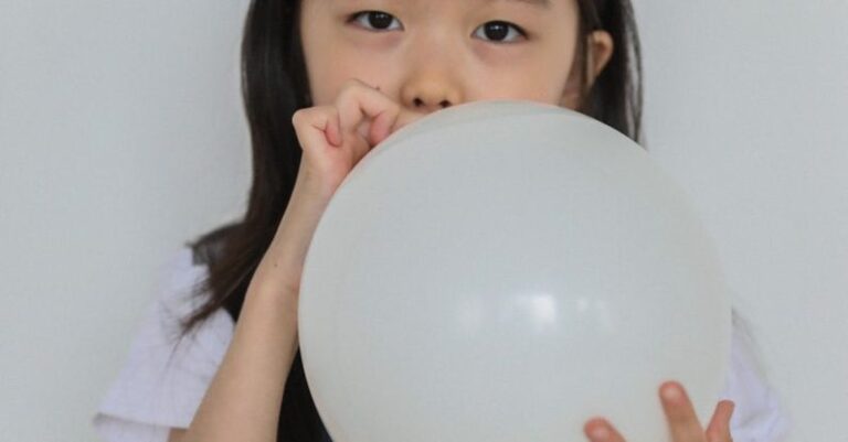 Inflate Deflate - Adorable little Asian girl with long dark hair in casual clothes inflating balloon and looking at camera against white background
