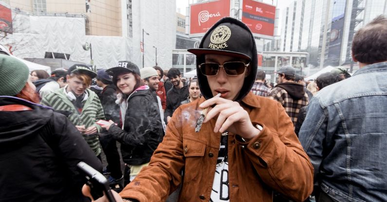 Joint Mobility - Young man in hat with hood standing on crowded street with smartphone in hand while smoking joint in city center