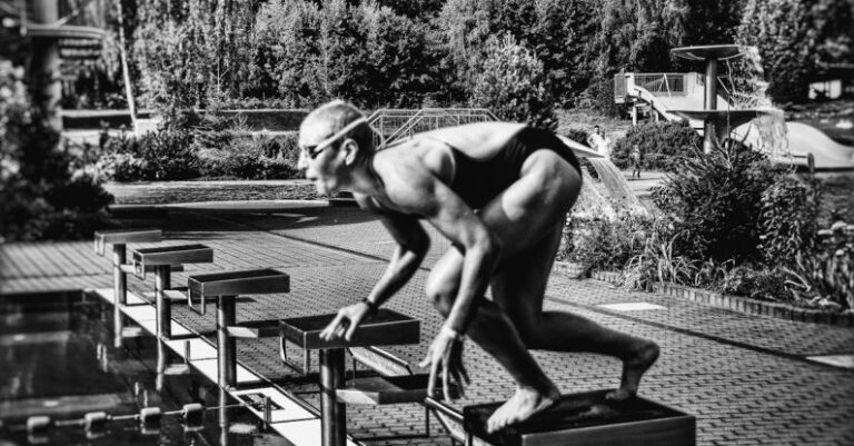 UV-Blocking Swimwear - Black and white side view full body sporty swimmer in swimming suit and goggles standing on block in track start position preparing to dive in outside pool