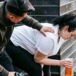 Hydrated - Sportsman clapping shoulder of plump tired female trainee sitting near net fence with bottle of water after training in autumn park