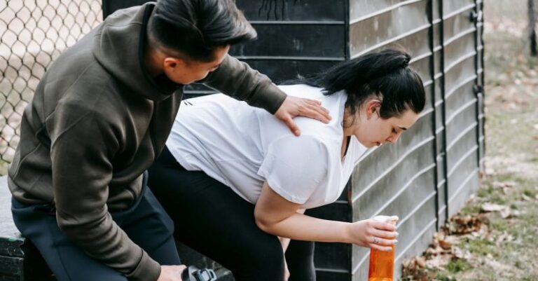 Hydrated - Sportsman clapping shoulder of plump tired female trainee sitting near net fence with bottle of water after training in autumn park