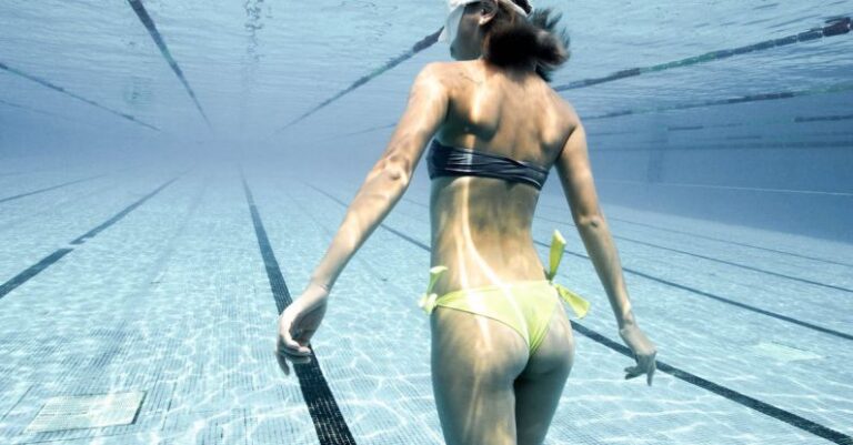 Night-Time Pool Activities - Back view of anonymous female in swimsuit and diving mask swimming underwater in clear water of swimming pool